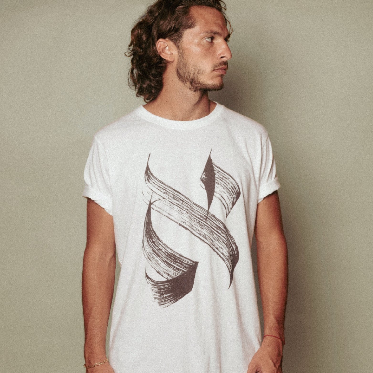 A man with wavy brown hair wears an Aleph white T-shirt made from premium cotton, featuring a bold abstract black brushstroke design. He stands against a plain light background, gazing to his right.
