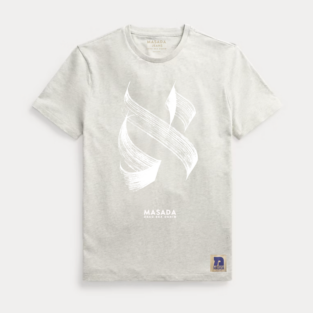 The Aleph white T-shirt features a premium cotton build with an abstract white brushstroke on the chest, MASADA DEAD SEA DEPTH text below, and a blue and gold patch at the hem—a nod to modern Israeli culture.