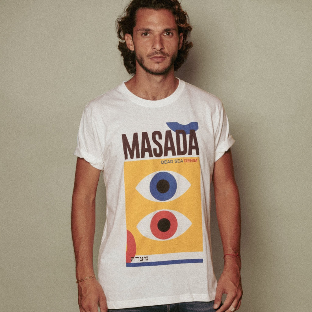 A man with wavy hair and a beard wears the HAYIN T-Shirt, a premium cotton tee from Masada Jeans featuring abstract eye designs in blue, red, and yellow with "MASADA DEAD SEA DENIM" printed above the graphic.
