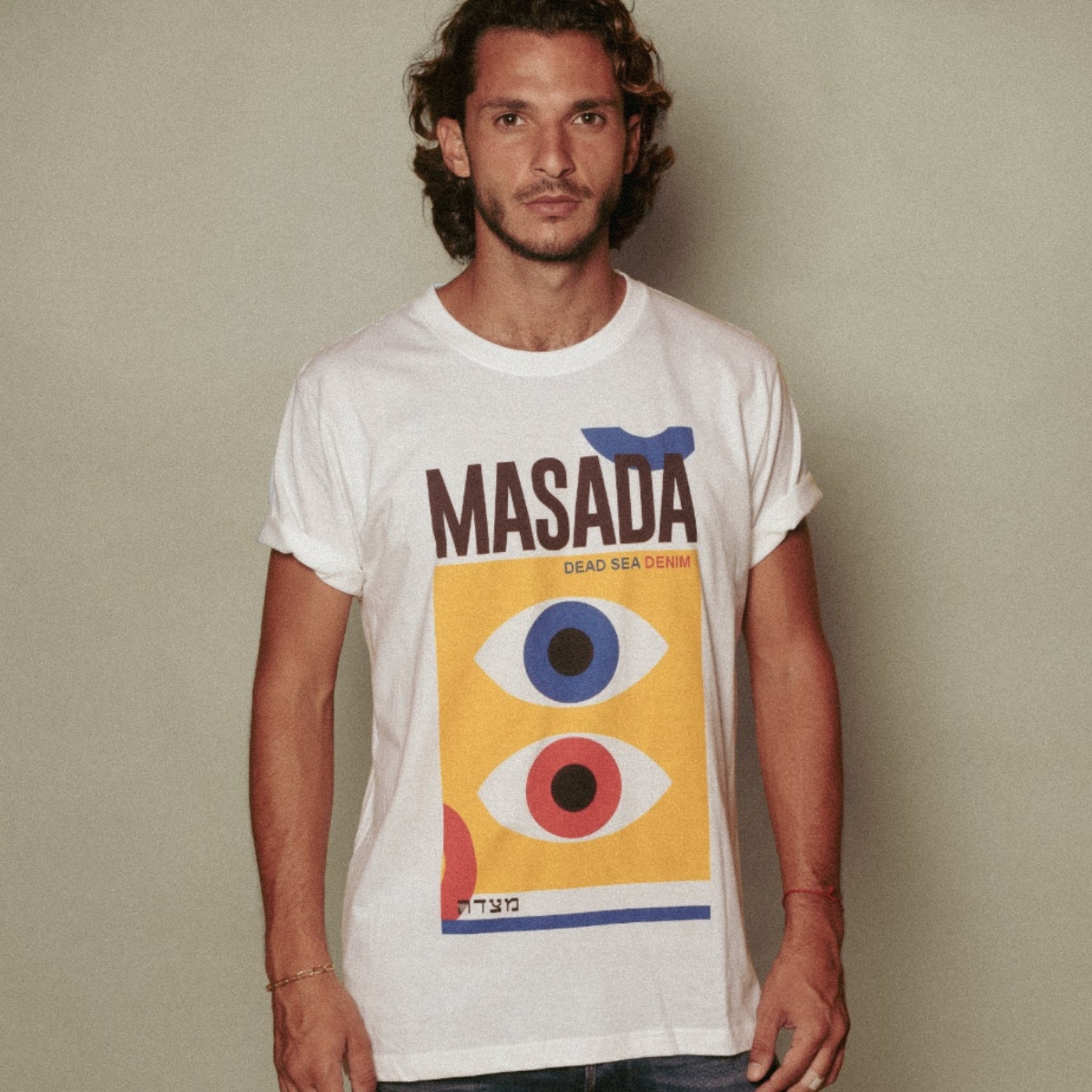 A man with wavy hair and a beard wears the HAYIN T-Shirt, a premium cotton tee from Masada Jeans featuring abstract eye designs in blue, red, and yellow with "MASADA DEAD SEA DENIM" printed above the graphic.